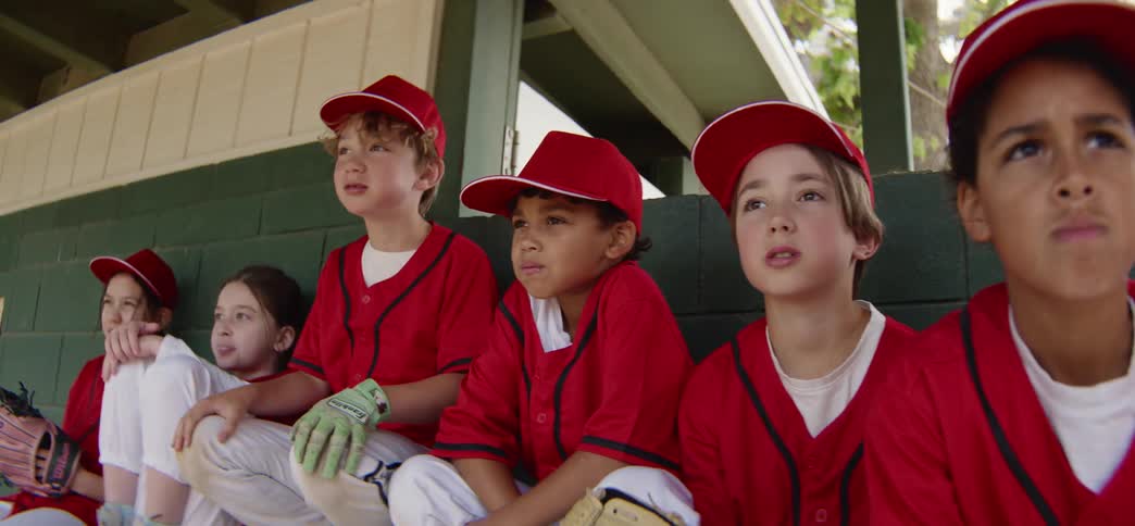 A diverse little league team watches their baseball game anxiously from the dugout