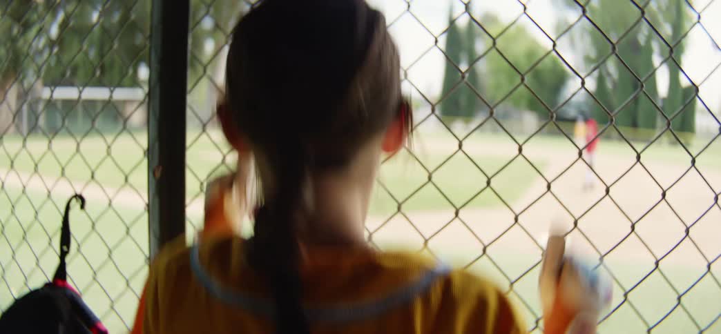A smiling little league player in a yellow jersey excitedly watches the game from behind the dugout fence