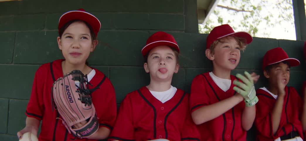 A diverse little league team in bright red uniforms enthusiastically cheers from the dugout bench in slow motion