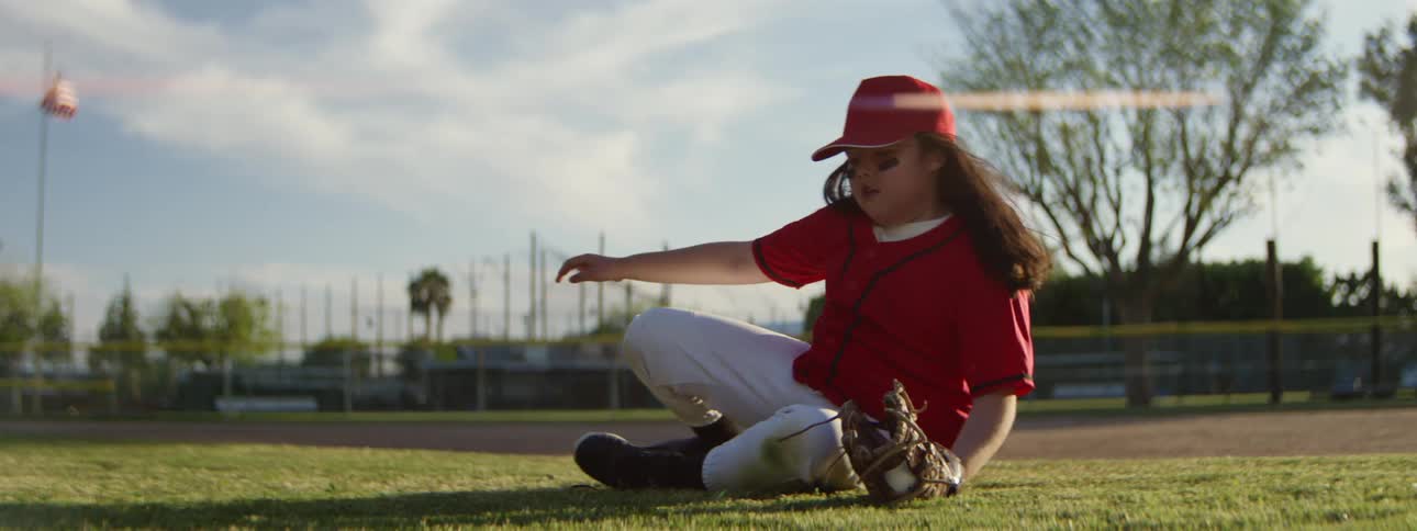 A determined young girl in a baseball uniform dives to catch a ball in the outfield in slow motion