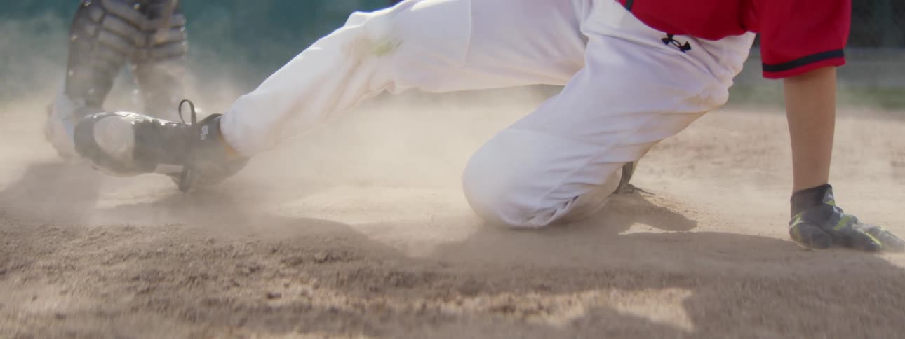 A baseball player slides safely into home plate in slow motion, kicking up a large cloud of dust