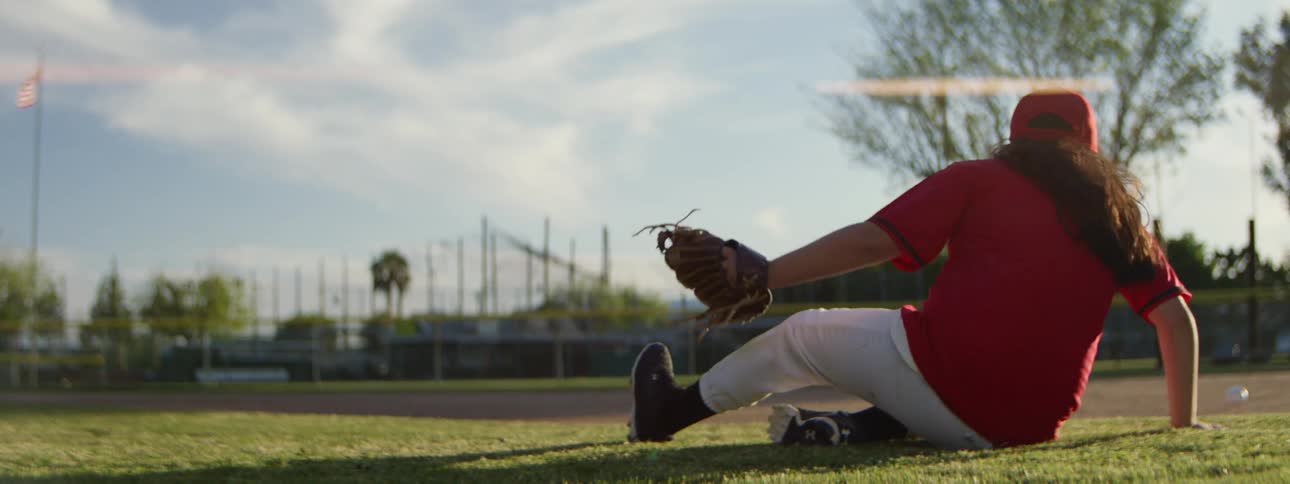 Slow motion shot of a young girl in a red baseball uniform diving for a grounder and missing the catch on a sunny field
