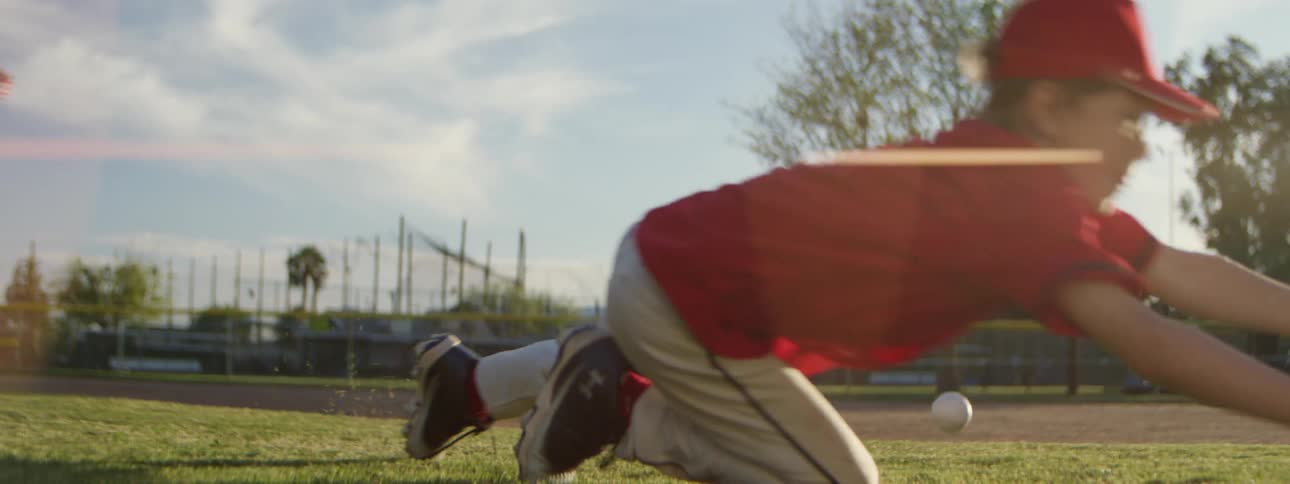 A young baseball player dives for a ground ball and misses the catch in an impressive slow-motion shot on a sunny day