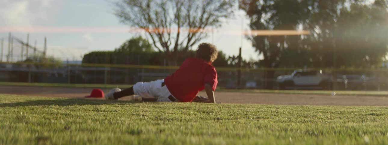 A young baseball player in a red uniform dives for a ground ball in slow motion, missing the catch and rolling onto his back on the sunlit field
