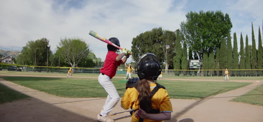 A young batter hits the ball in slow motion as seen from over the catcher's shoulder during a sunny little league baseball game