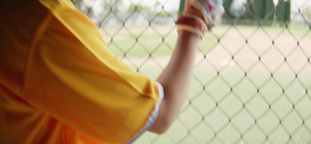 A young girl in a yellow jersey puts on her batting gloves in the dugout before looking out at the baseball field with a smile
