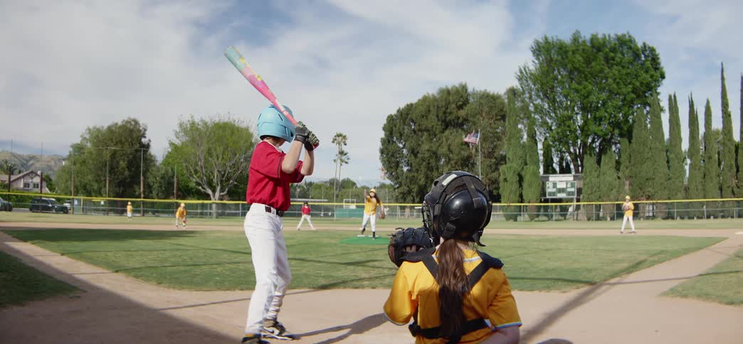 Slow-motion view from behind the catcher of a young batter hitting the ball during a little league baseball game