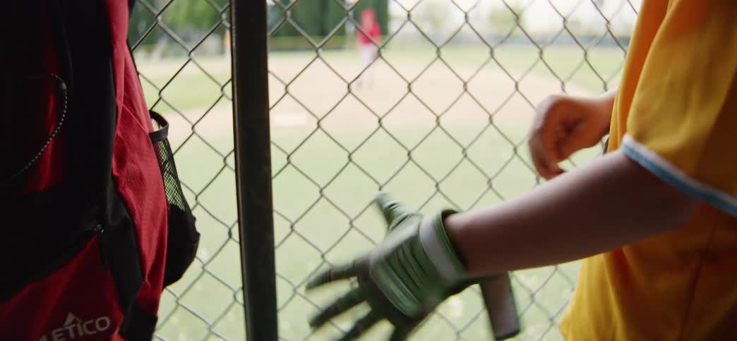 From the dugout, a young baseball player slowly puts on their batting glove, watching the game through a chain-link fence