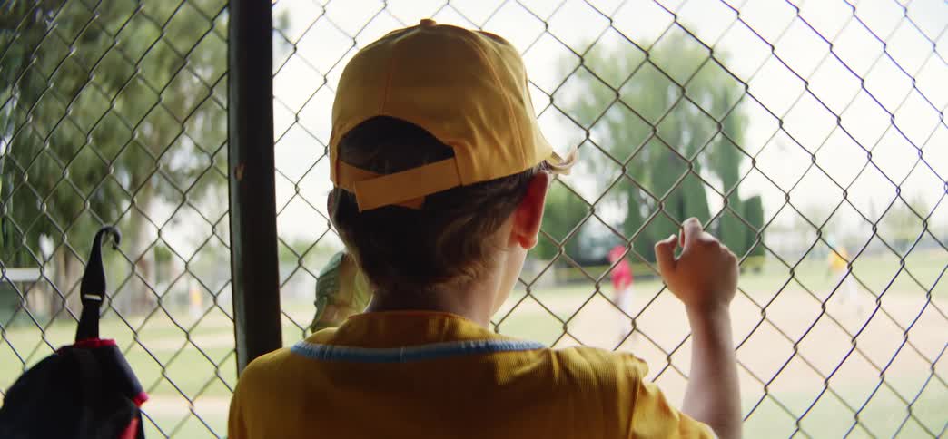A young little league player in a yellow uniform watches his team play from the dugout through a chain-link fence