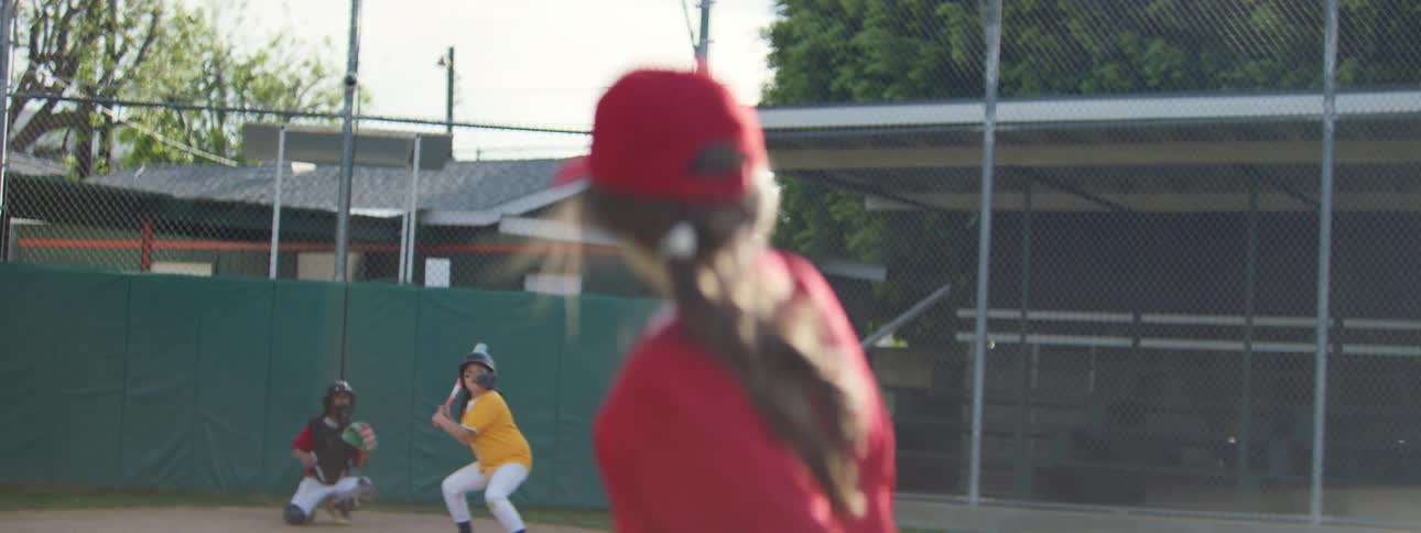 Slow motion over-the-shoulder view of a girl in a red uniform pitching a baseball to the batter during a game