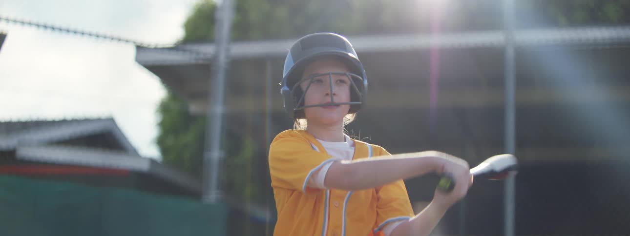 Determined young girl in a yellow uniform swings a baseball bat with a smile in slow motion on a sunny field