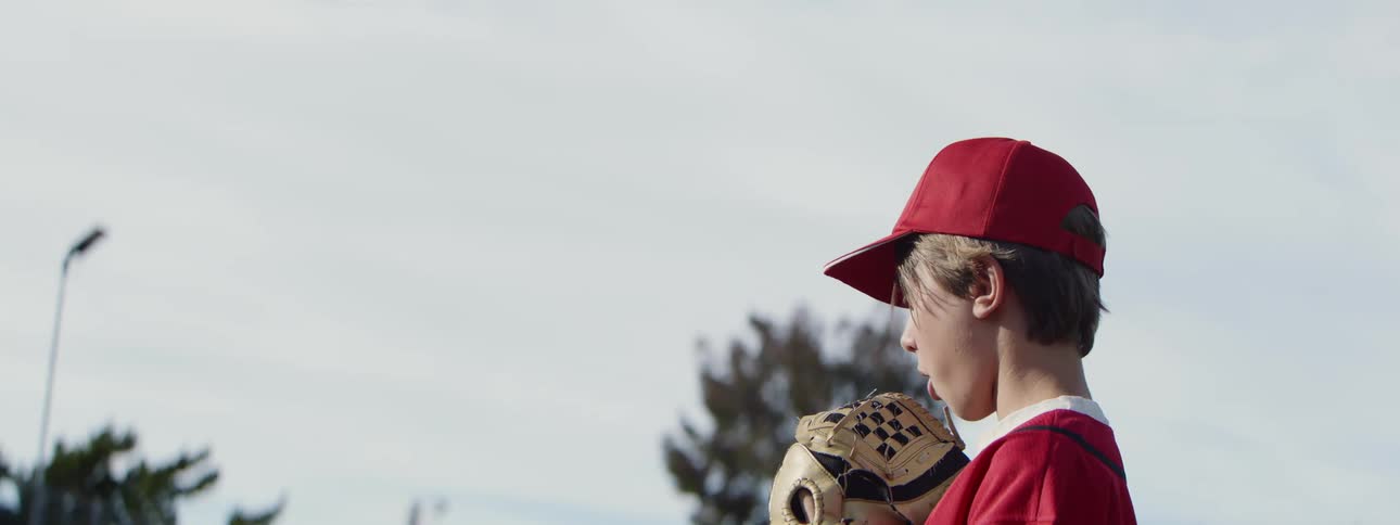 A young boy in a red uniform and baseball cap stands focused on a baseball field in slow motion