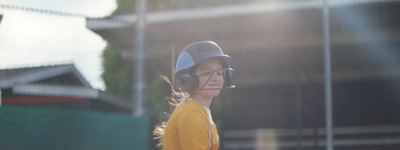 A joyful little girl in a baseball uniform swings her bat with a big smile in slow motion and runs off the field