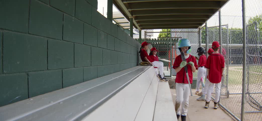 A young baseball player walks through the dugout in slow motion and sits on the bench, waiting for his at-bat