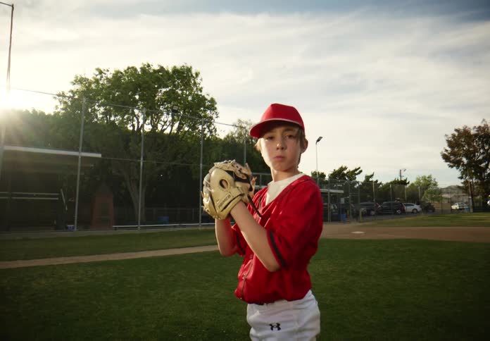 A determined young baseball pitcher in a red uniform winds up and throws a ball directly at the camera in slow motion