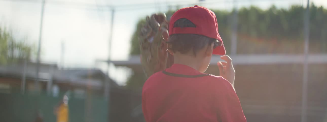 Slow motion shot from behind a young baseball player as he puts on his red cap on a sunny field