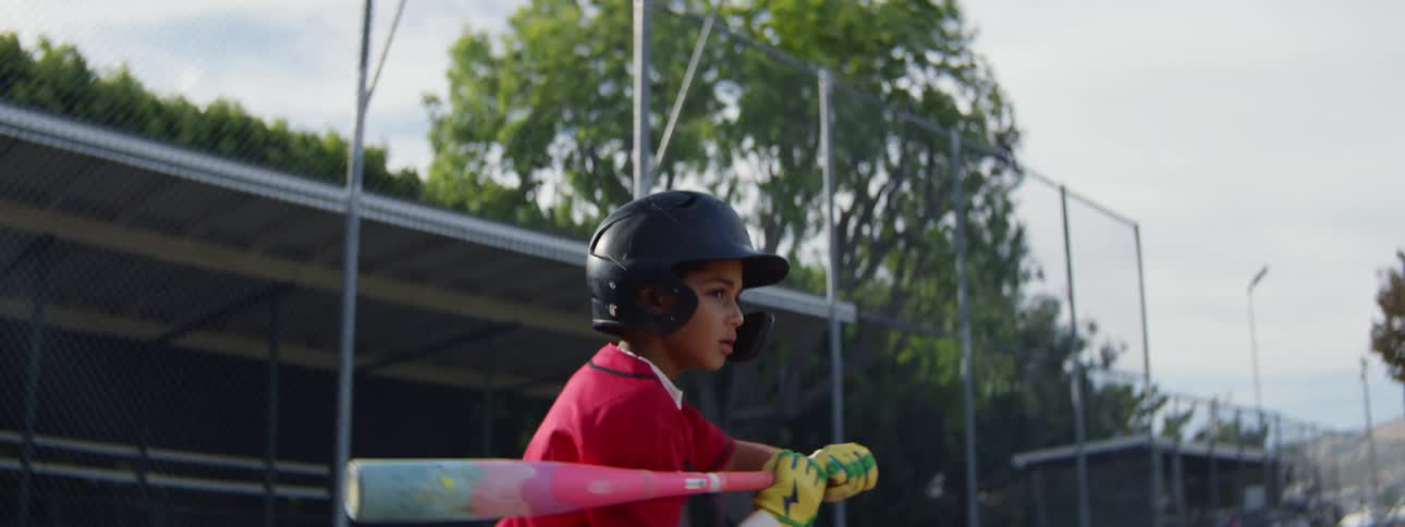 Slow motion view of a young boy in a baseball uniform and helmet swinging a bat and dropping it after the play