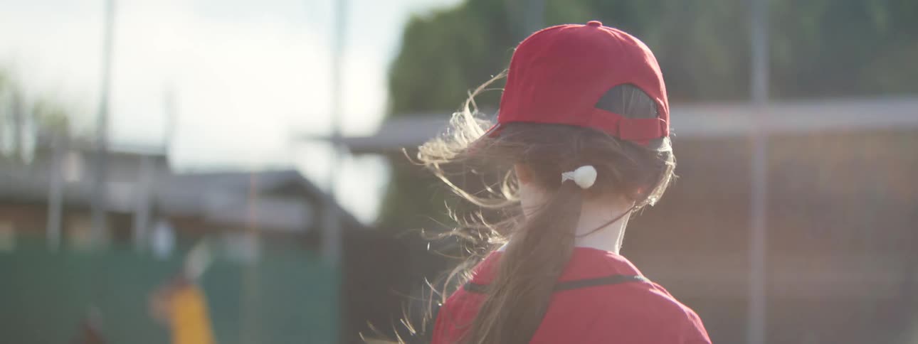 The wind blows through a young girl's ponytail in slow motion as she stands on a baseball field in the sun