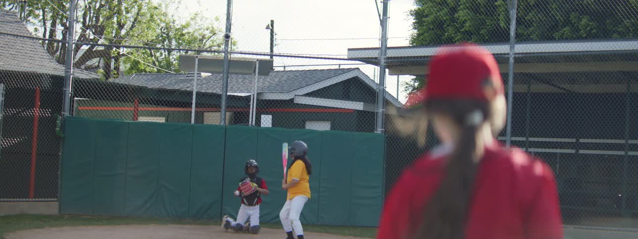 Slow motion shot from behind a young female pitcher as she throws a baseball to the batter and catcher during a game