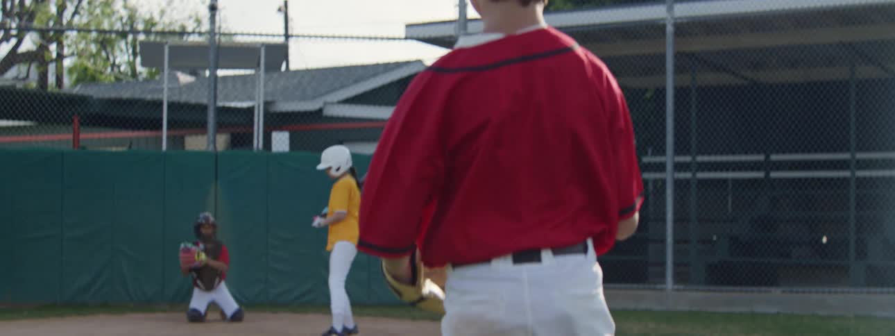 Slow motion over the shoulder view of a young boy in a red uniform pitching a baseball to a batter on a sunny day