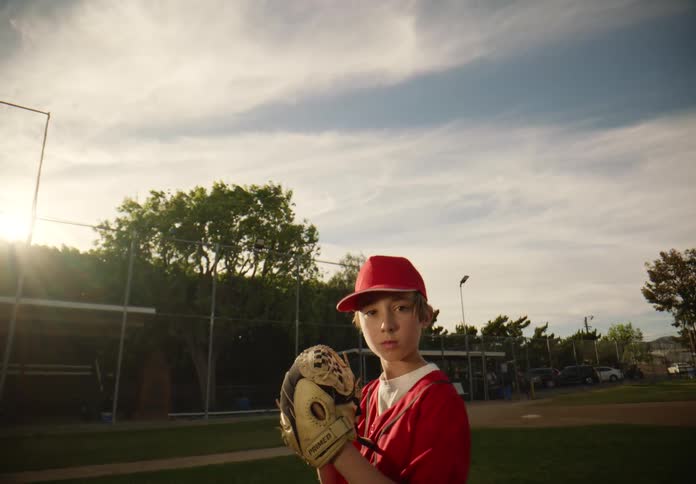 Point-of-view shot of a young boy in a red uniform pitching a baseball towards the camera in slow motion on a sunny field