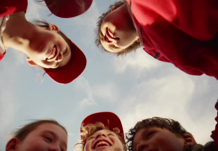 A low-angle view of a diverse group of happy children in a sports team huddle, smiling down at the camera under a sunny sky