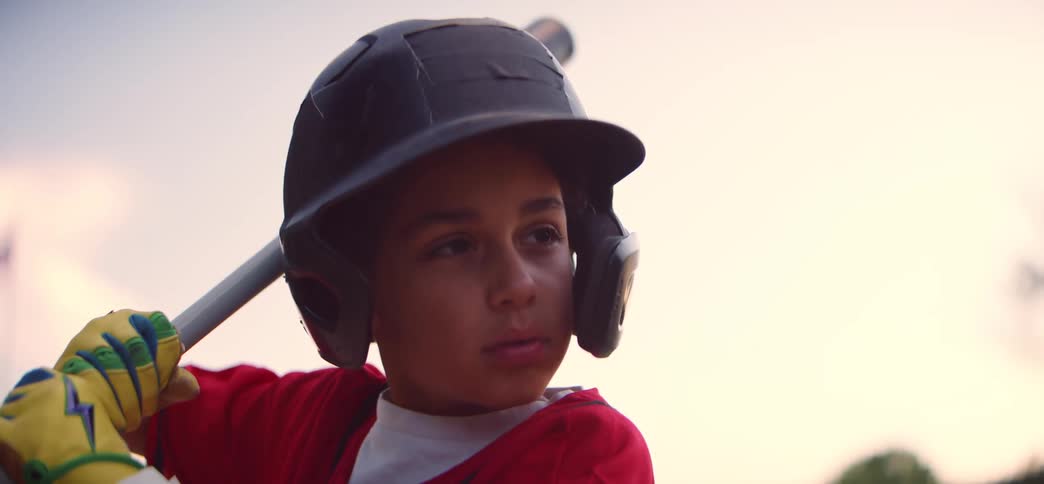 A determined young baseball player in a red uniform and batting helmet concentrates intently as he waits for the pitch