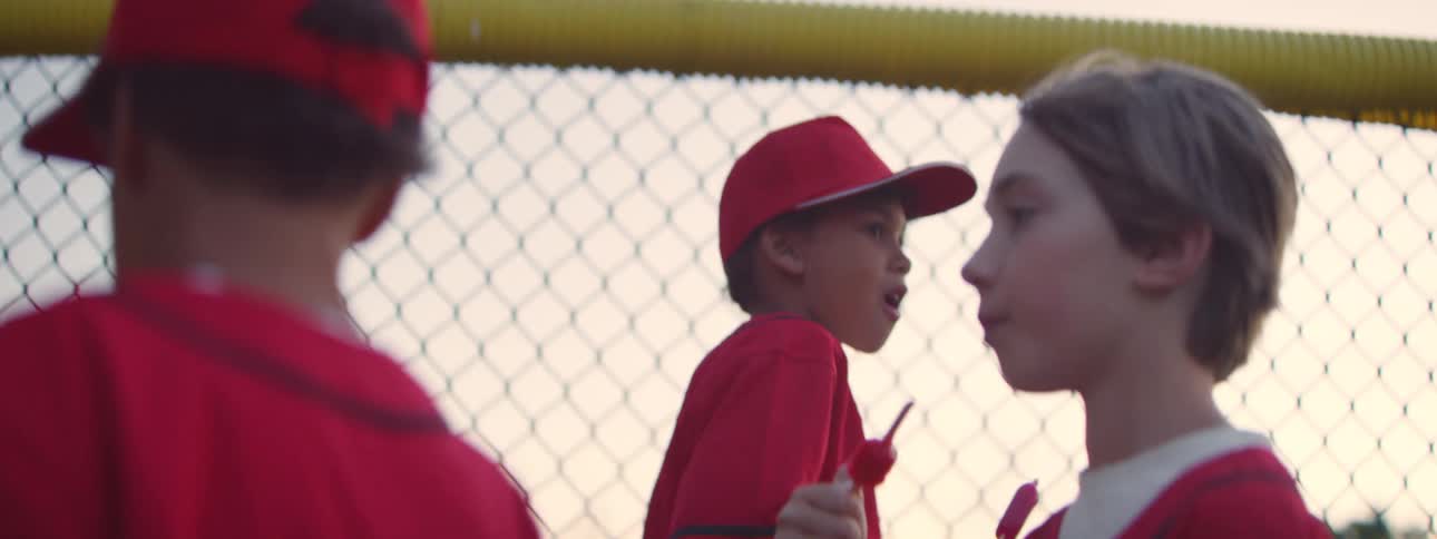 A slow-motion close-up of young baseball players in red uniforms eating lollipops by a fence