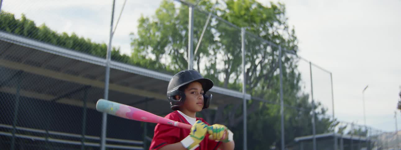 A young baseball player in full gear swings his colorful bat with determination and starts running the bases in slow motion