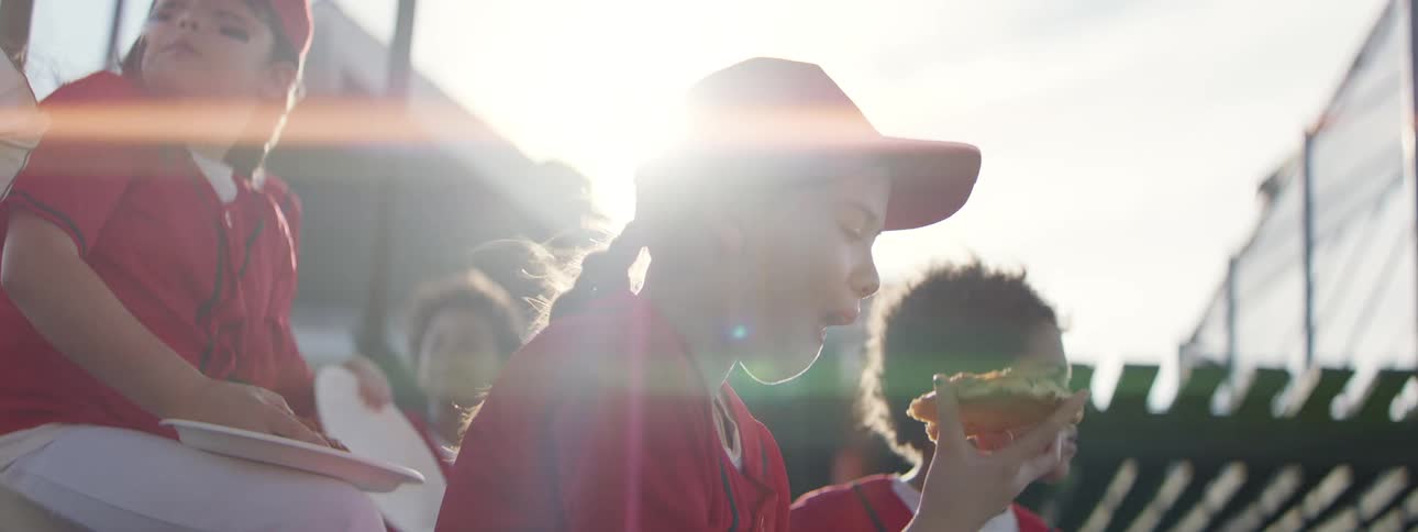 A young girl in a red baseball uniform eats a slice of pizza in slow motion while sitting on the bleachers with her teammates