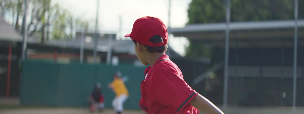 From over the shoulder of the pitcher, a young girl at bat swings and misses the ball in slow motion during a little league game