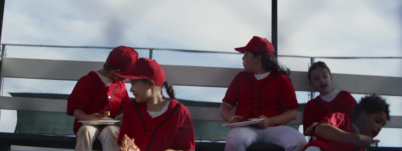 A diverse youth baseball team in matching red uniforms celebrates with pizza on the bleachers in slow motion