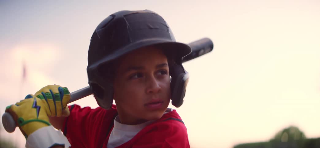 An intensely focused young baseball player in a red uniform holds his bat, ready to swing during a game at sunset
