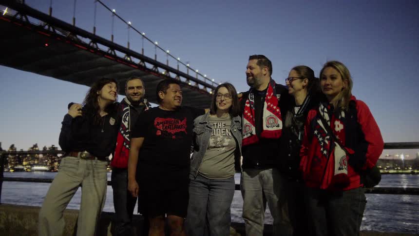A diverse group of American soccer fans smiles for a photo under an illuminated city bridge at dusk