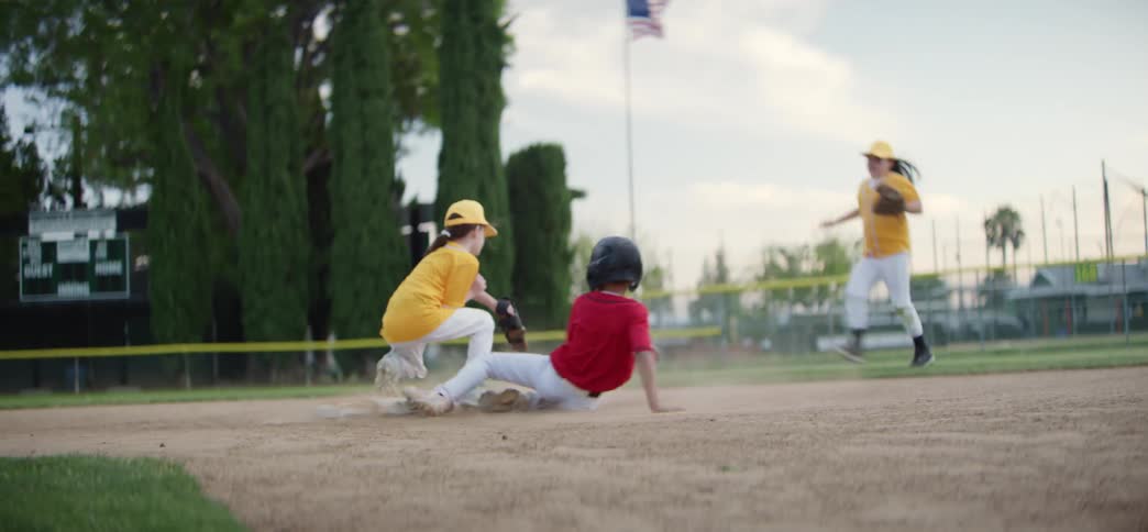 In a dramatic slow-motion sequence, a young baseball player in a red jersey slides safely into a base during a little league game