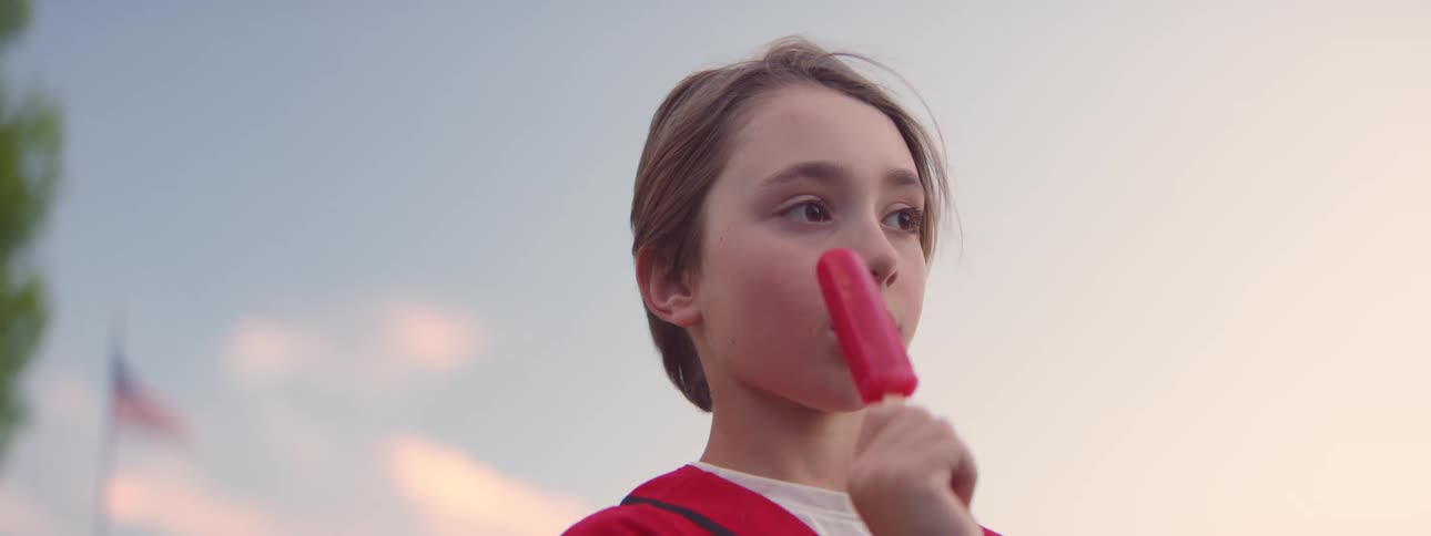 A young boy in a baseball jersey savors a red popsicle outdoors on a summer evening in slow motion