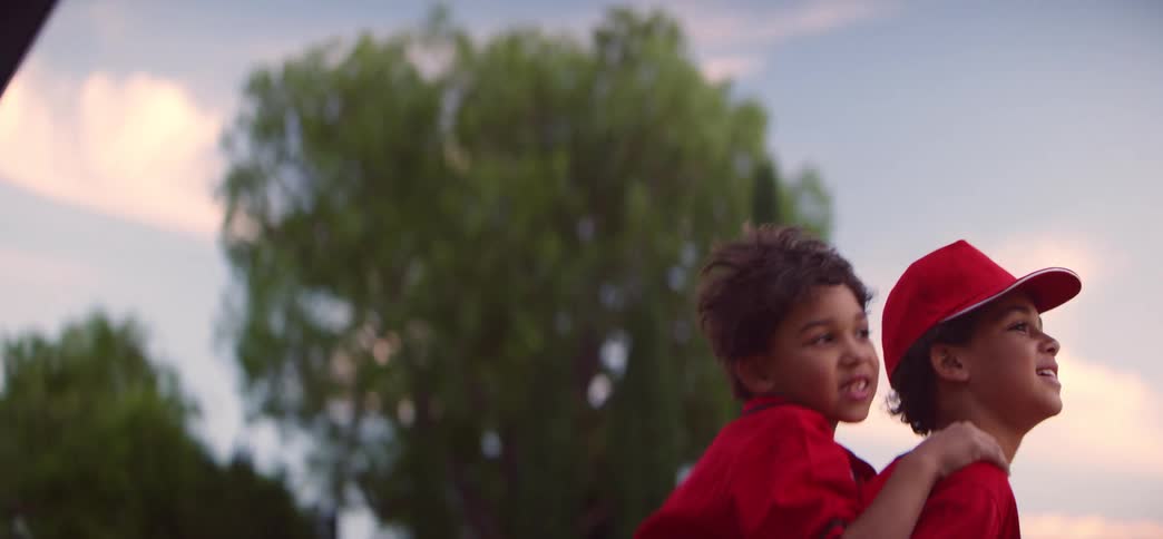 An older brother gives his smiling little brother a piggyback ride across a baseball field in slow motion at sunset