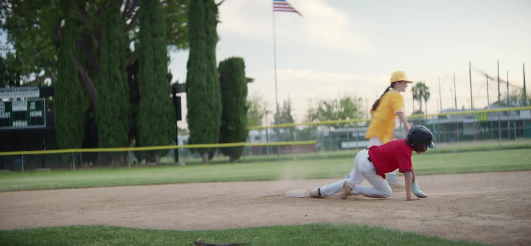 A determined young baseball player in a red shirt sprints and slides safely onto the base during a little league game in slow motion