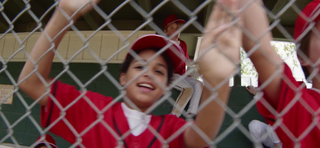 Excited young little league baseball players in red uniforms cheer enthusiastically for their teammates from behind the dugout fence