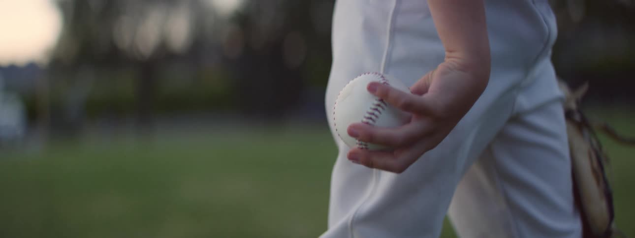 A young baseball player's hand holds a baseball on a grassy field in the evening light