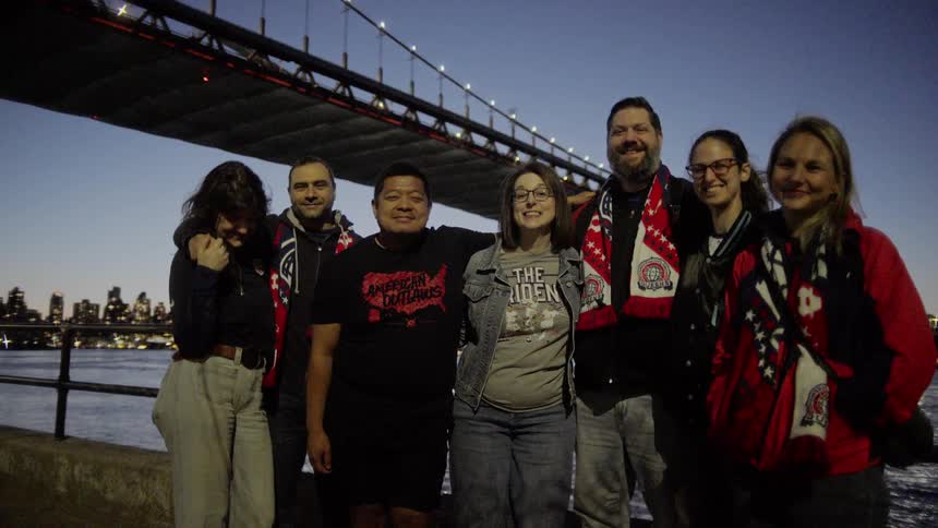 A group of happy friends wearing sports scarves pose for a photo on a city waterfront at dusk under a large illuminated bridge