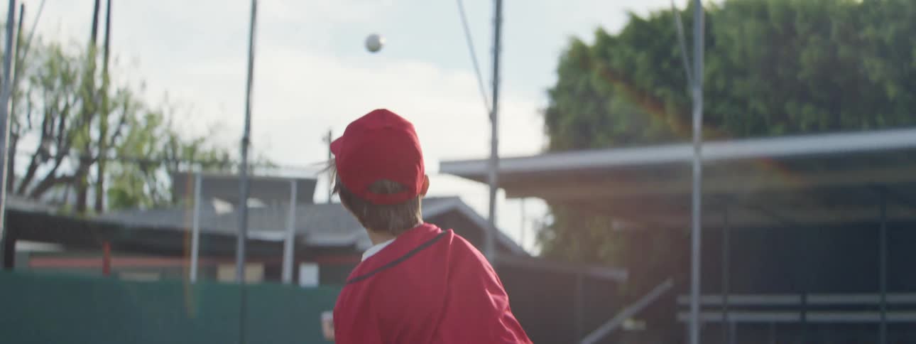 A young boy in a red uniform pitches a baseball in slow motion during a sunny Little League game