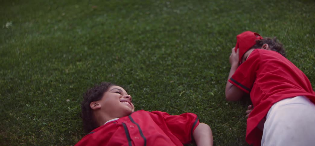 A high-angle slow-motion shot of two young baseball players in red jerseys lying on the grass and smiling