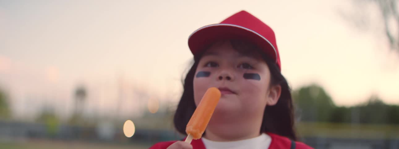 Slow motion close-up of a happy young baseball player in a red uniform enjoying a refreshing orange popsicle after a game at dusk