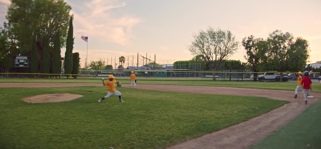 A young batter in a red shirt runs for home plate in slow motion as the pitcher fields the ball and makes a throw during a little league game