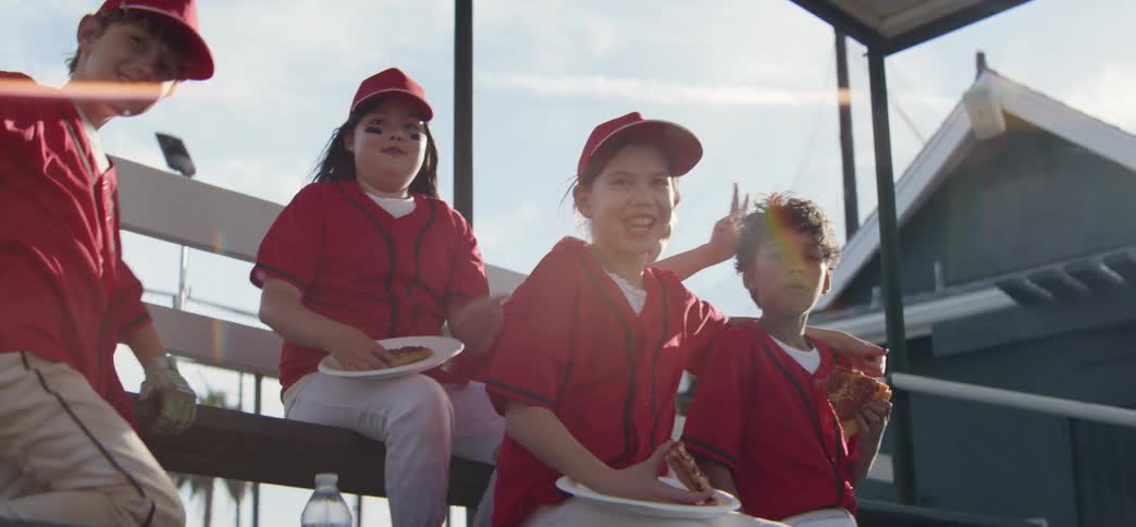 A diverse little league team in red uniforms laughs and eats celebratory pizza on the dugout bench in the bright sunshine