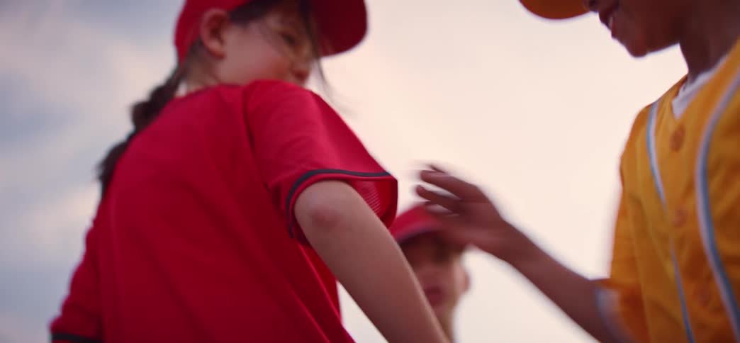 A low-angle shot captures a diverse children's baseball team putting their hands together in a huddle for a pre-game cheer