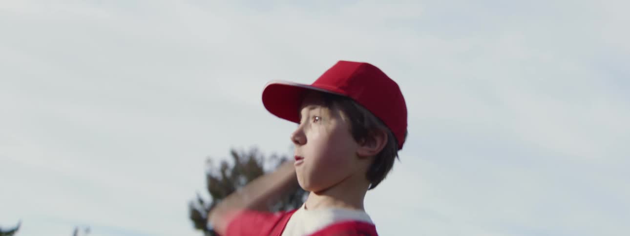 A determined young boy in a red baseball uniform winds up and pitches in slow motion on a sunny day