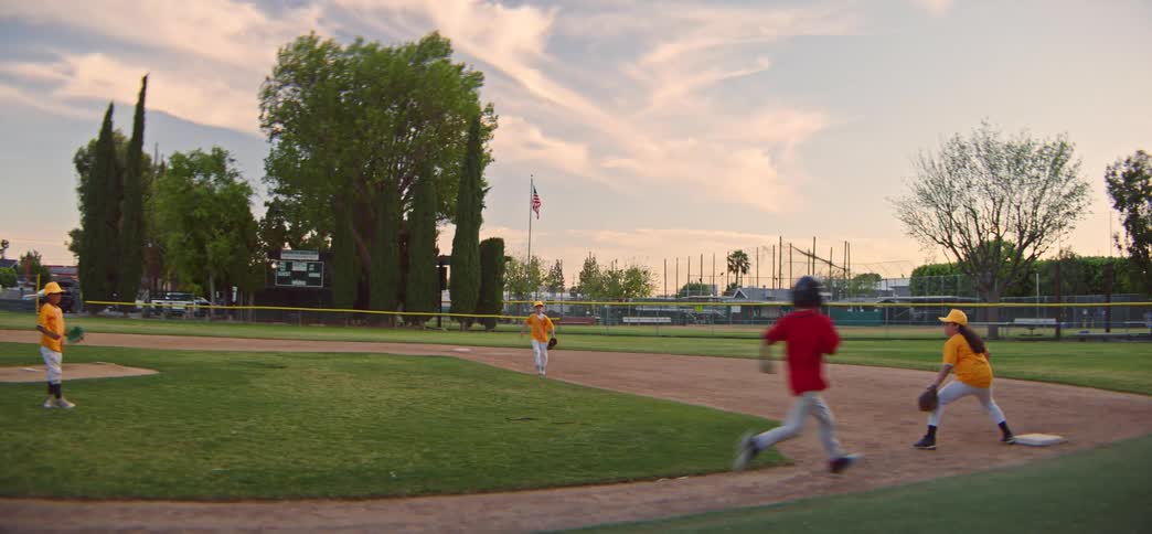 A young boy in a red uniform runs to first base as his teammates field the ball during a little league game on a sunny evening