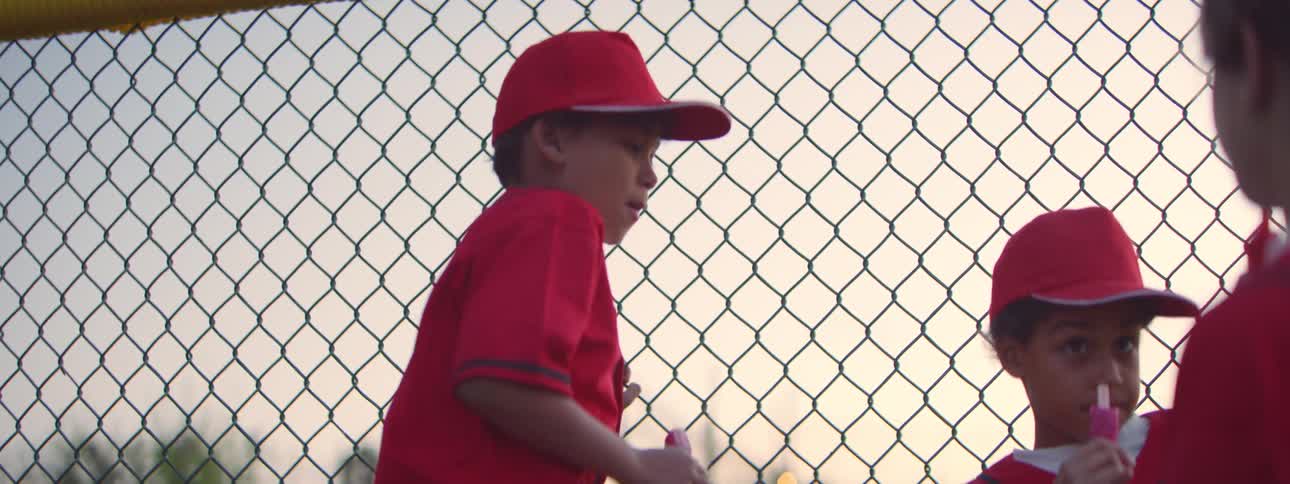 A young baseball player in a red uniform eats a pink popsicle by a fence as the shot slowly comes into focus